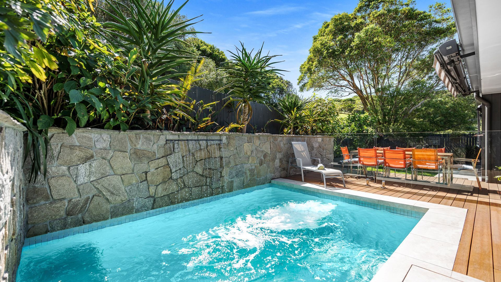 A swimming pool with a stone wall, surrounded by lush greenery and a wooden deck with outdoor furniture.