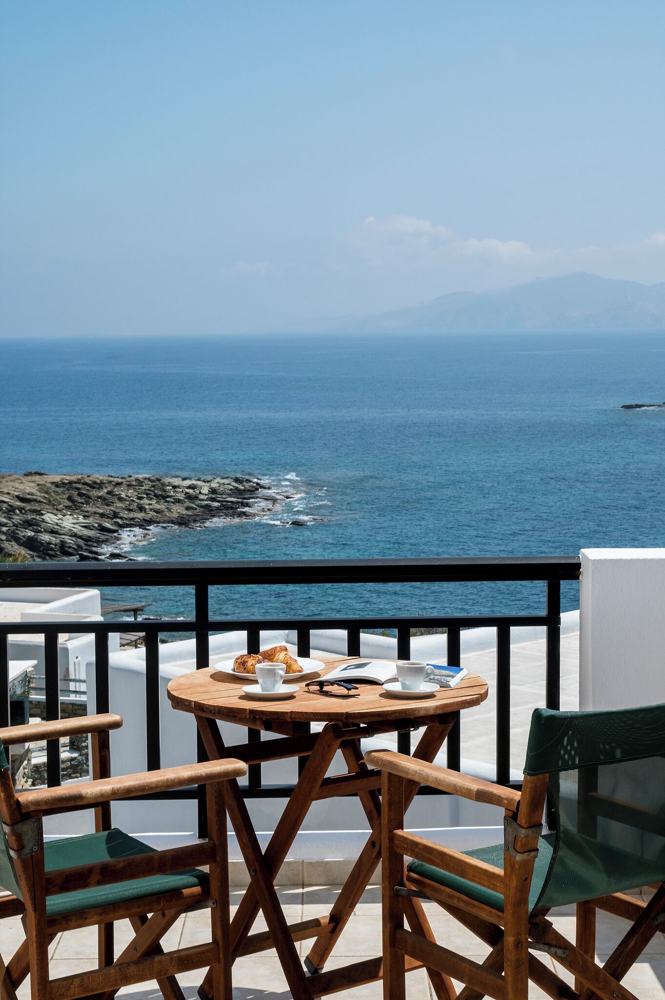 A balcony with a wooden table and chairs, overlooking a rocky coastline and the sea.