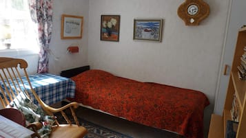 A small, cozy room with a single bed, a wooden chair, a table with a checkered tablecloth, and a bookshelf.