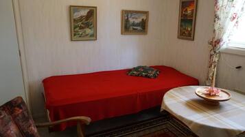 Una habitación con una cama individual, una silla, una mesita con una flor y dos cuadros en la pared.