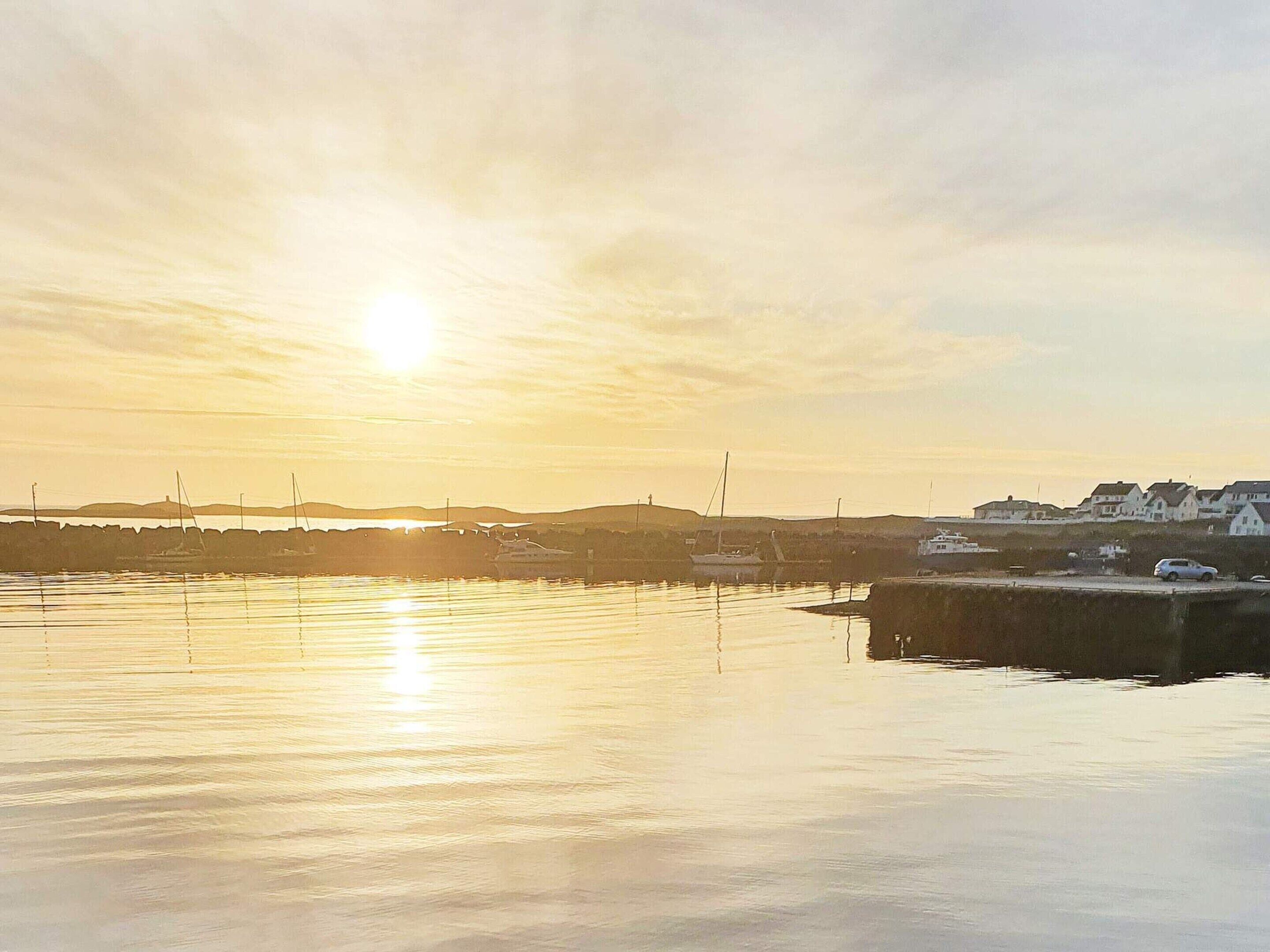 A coastal marina with boats docked, calm water reflecting the sunset, and buildings in the background.