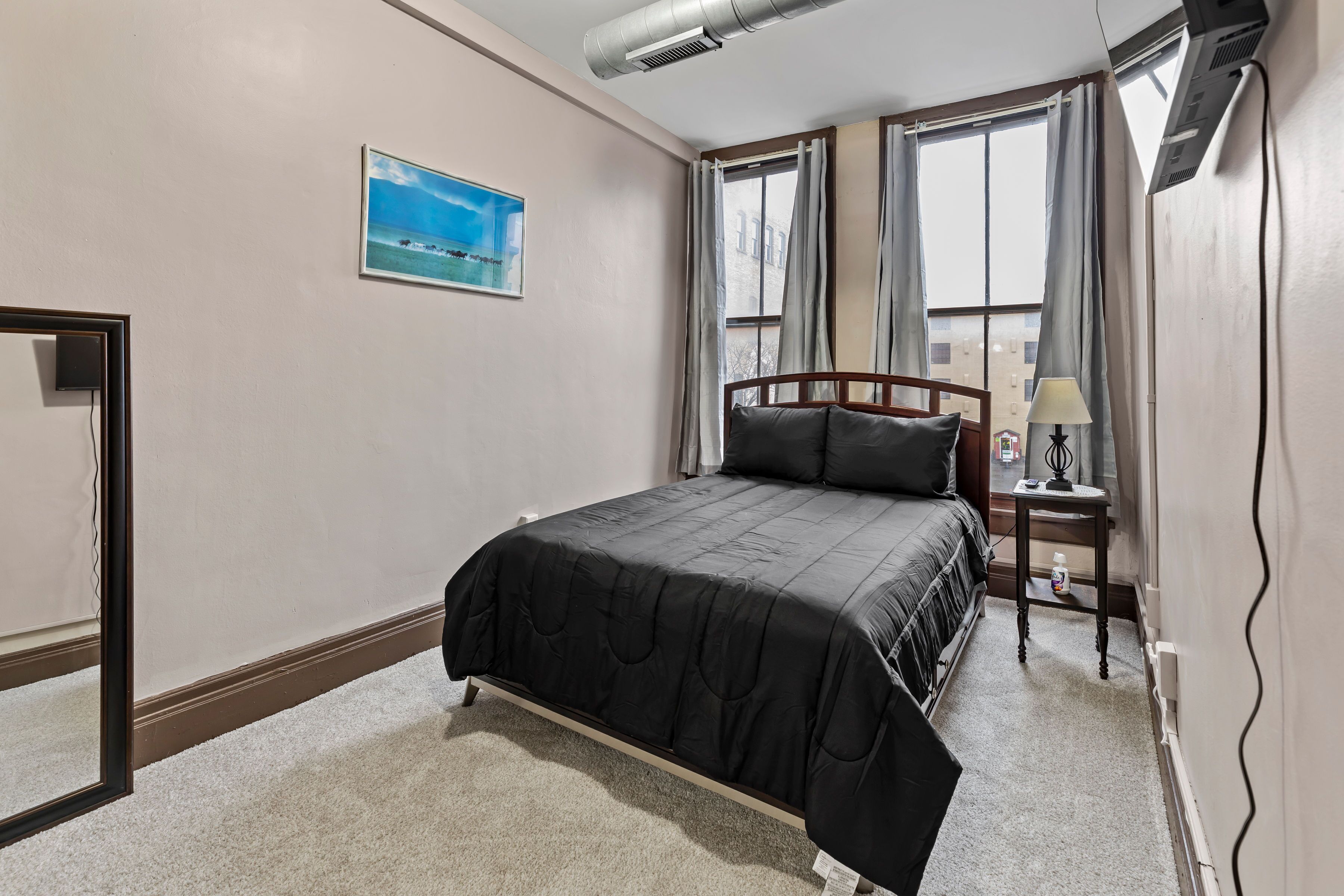 Une chambre d’hôtel avec un lit, une table de chevet, une lampe, une fenêtre avec des rideaux et un tableau au mur.