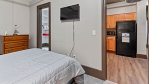 Une chambre à coucher avec un lit, une commode, un téléviseur fixé au mur et un coin cuisine visible à travers une porte ouverte.