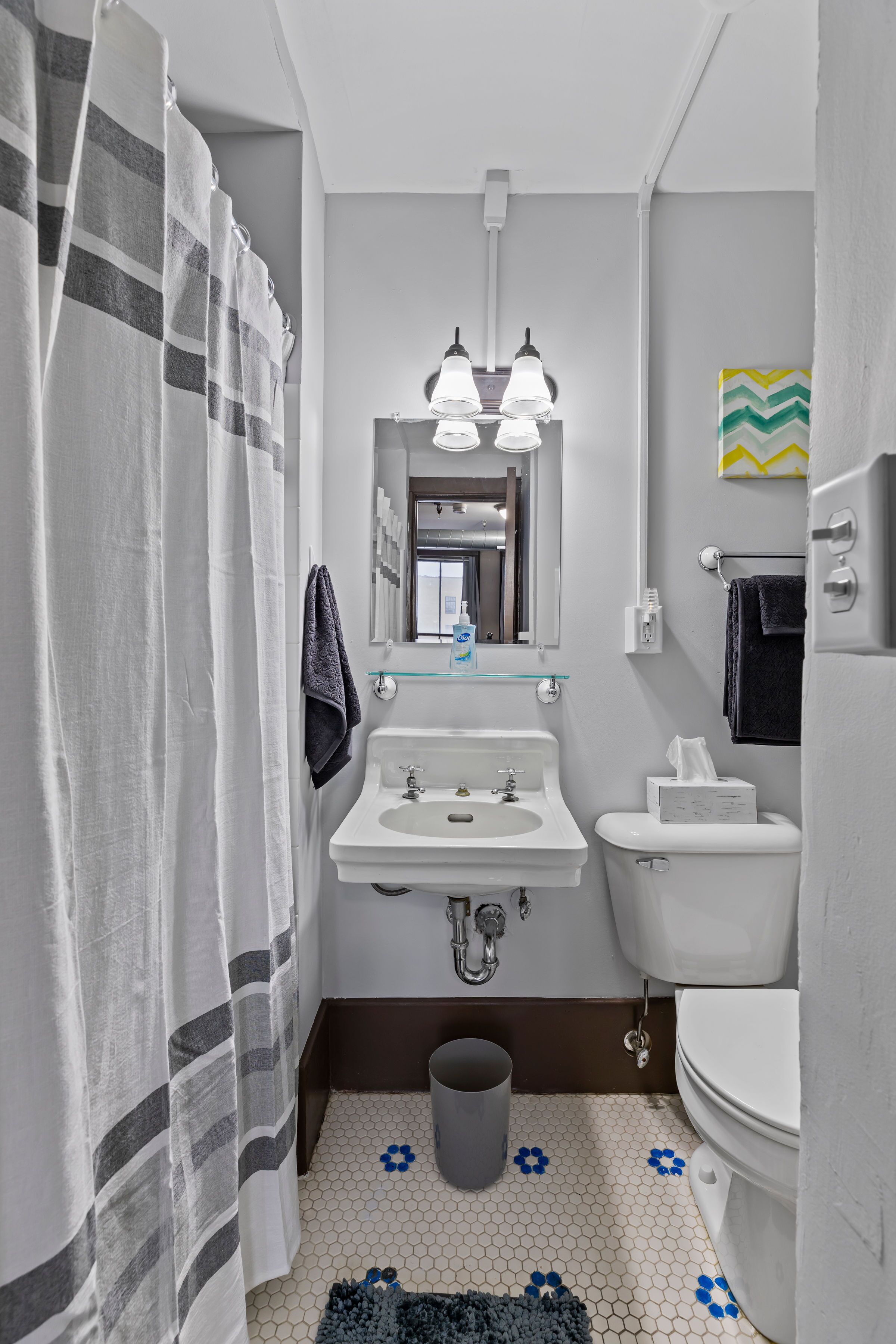 Une salle de bain avec un lavabo, des toilettes et un rideau de douche, tous de couleur blanche. Un miroir est placé au-dessus du lavabo, et il y a un porte-serviettes.