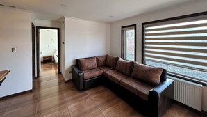 A modern living room with a sectional sofa, wooden flooring, and large windows with blinds. - Alto Balmaceda Apartamentos (Natales)