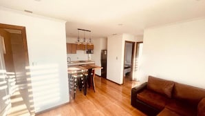 A spacious living area with a wooden floor, a brown leather sofa, and a kitchen in the background. - Alto Balmaceda Apartamentos (Natales)