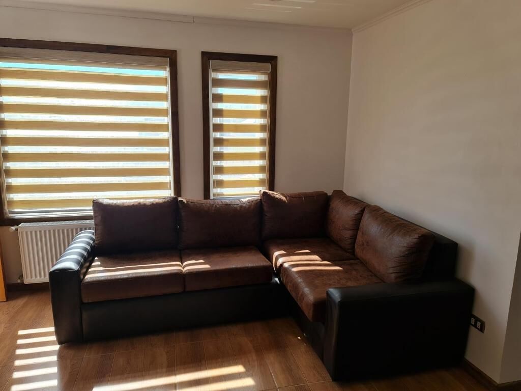 A living room with a brown leather sectional sofa, two windows with blinds, and a radiator.