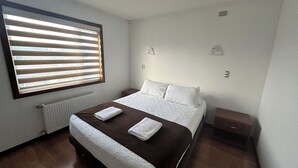 A bedroom with a large bed, a window with blinds, a radiator, and a nightstand with a lamp. - Alto Balmaceda Apartamentos (Natales)