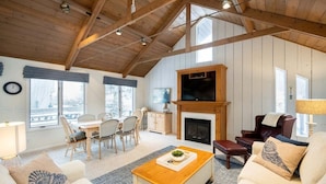 A spacious living room with a high ceiling, wooden beams, a fireplace, a television, and a dining area with a view of the outdoors. - Updated cottages steps from Lake Michigan beach (West Olive)