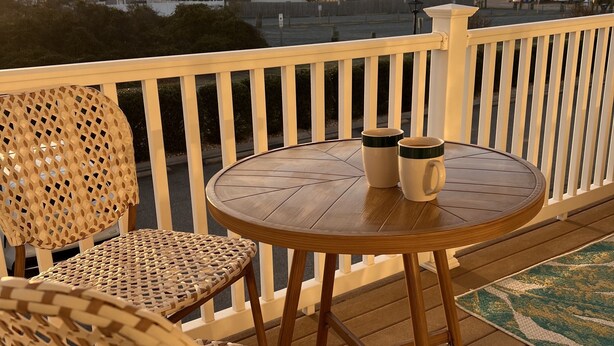 A wooden deck with a round table and two wicker chairs, a white railing, and a view of buildings in the background. - Second Row Condo with Ocean Views, Pool, and Hot tub (Oak Island)