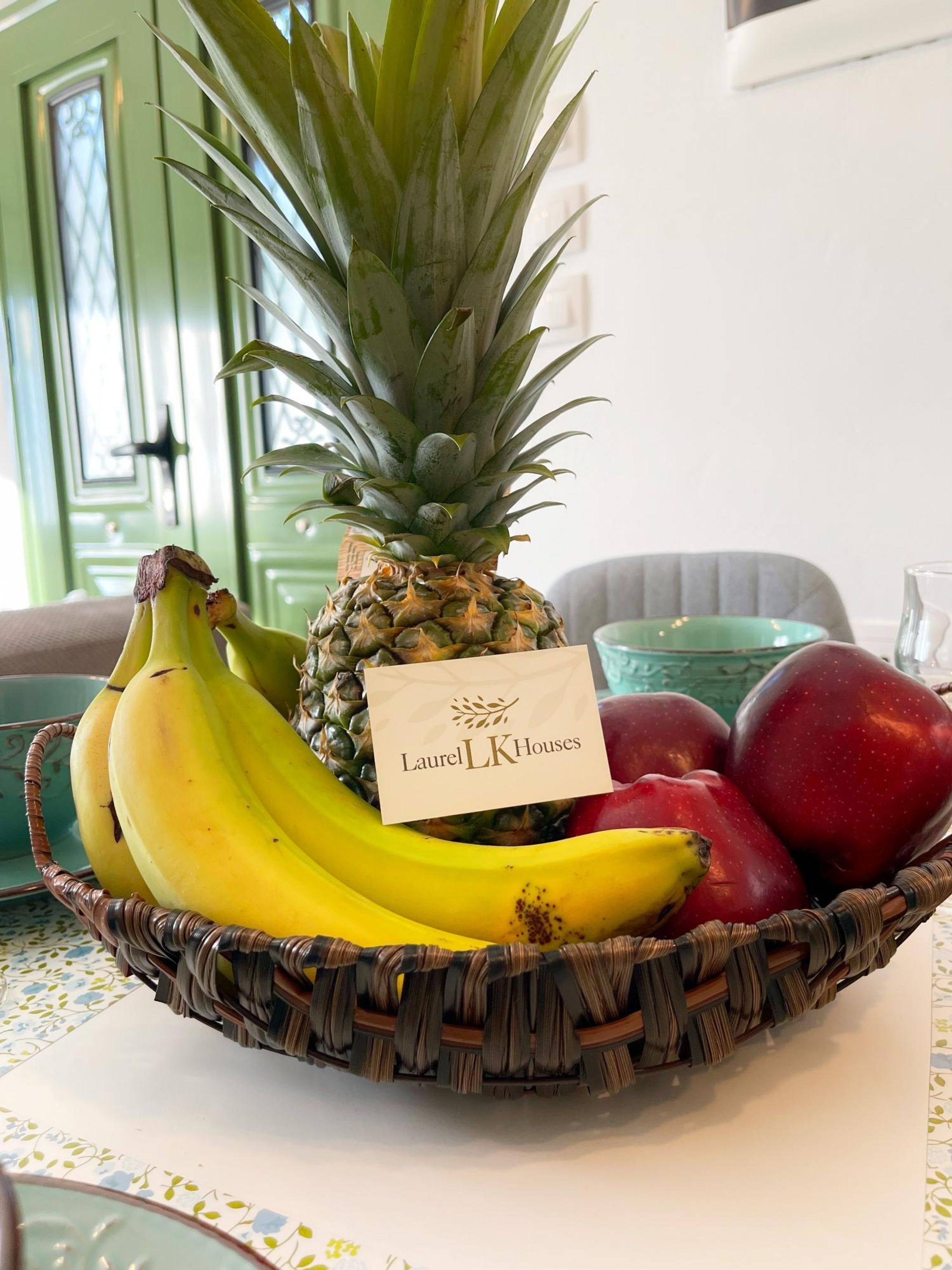 Un panier en osier rempli de fruits – des bananes, un ananas et des pommes rouges – est posé sur une table.