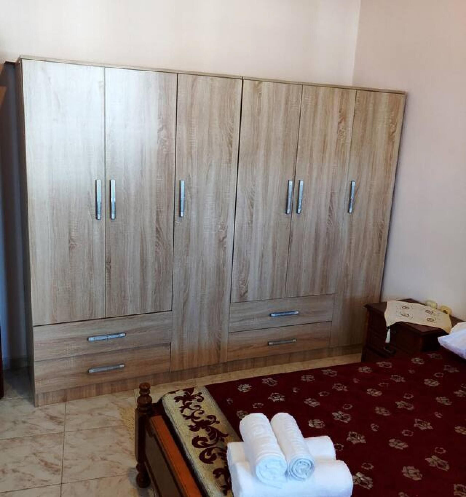 A bedroom with a large wooden wardrobe, a bed with a red patterned bedspread, and a small bedside table with a lamp.