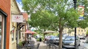 Un café al aire libre con mesas y sillas, un edificio de ladrillo y automóviles estacionados.