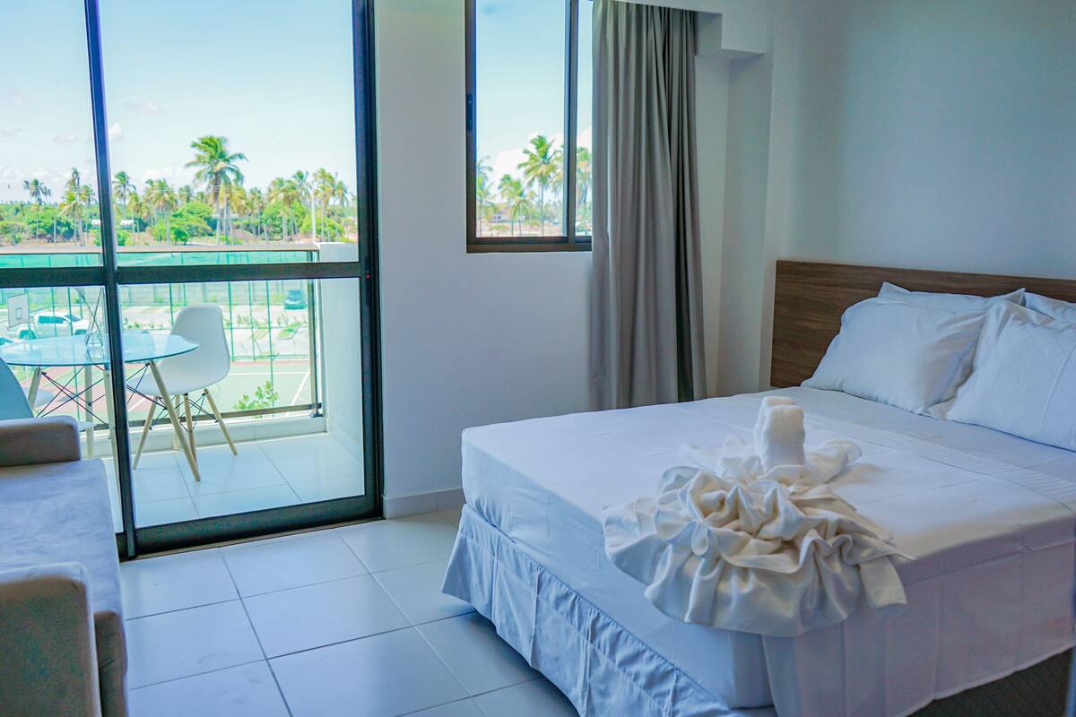 Habitación de hotel con cama, balcón con mesa y sillas, y vista a palmeras.