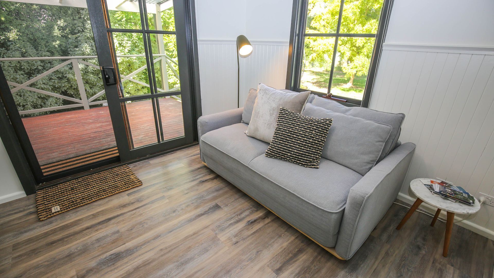 Un salon moderne avec un canapé gris, une lampe sur pied et une vue sur un jardin grâce à de grandes fenêtres.