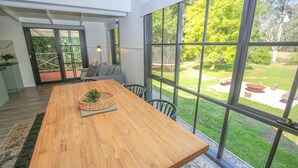 A spacious room with a large wooden table, a wicker side table, and a view of a garden through large windows. - LUMLEY ABODE : Bright (Bright)
