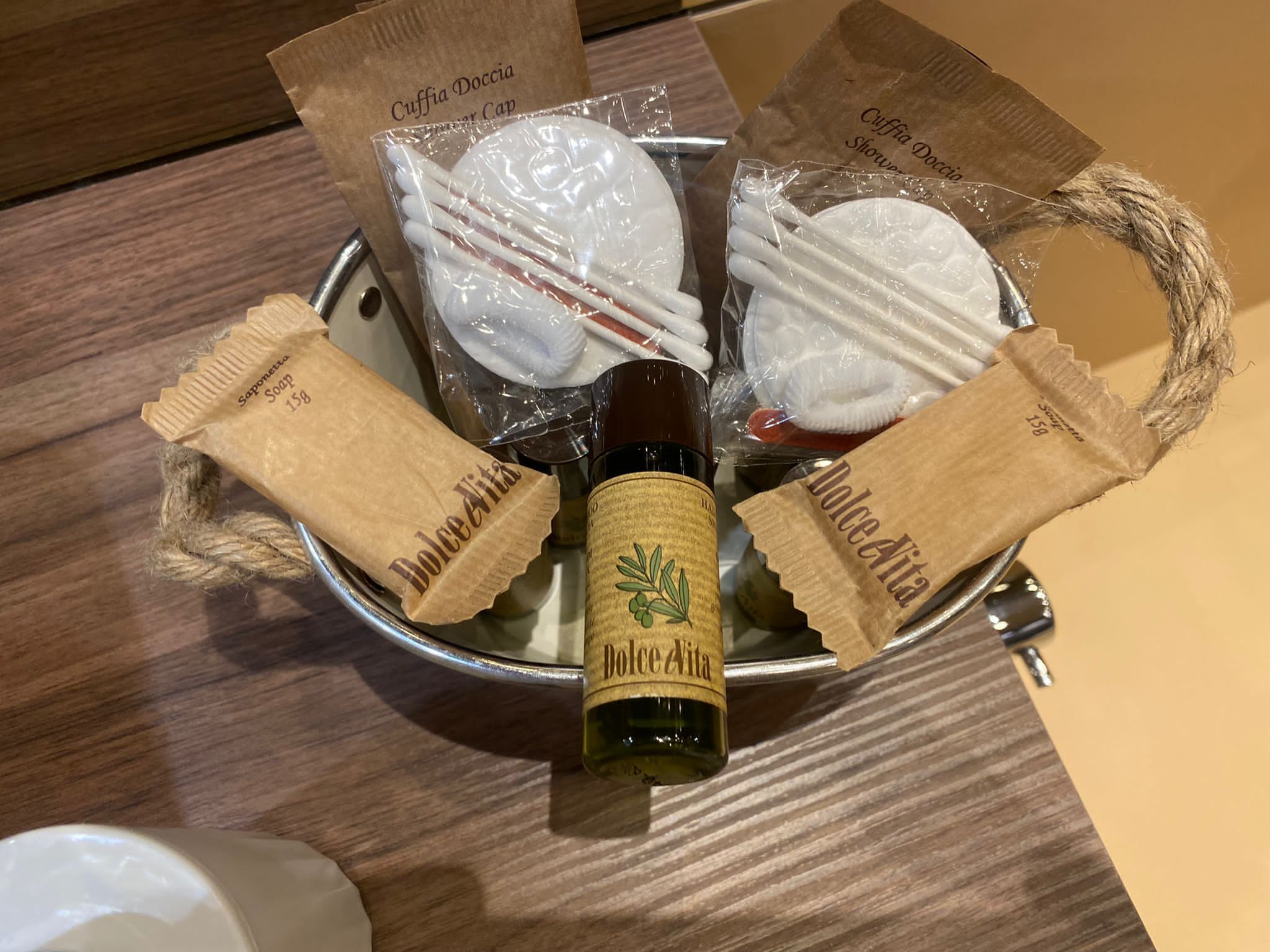 A metal basket containing a bottle of Dolce Aita wine, two bars of soap, and two packages of Cuffia Doccia shower cap.