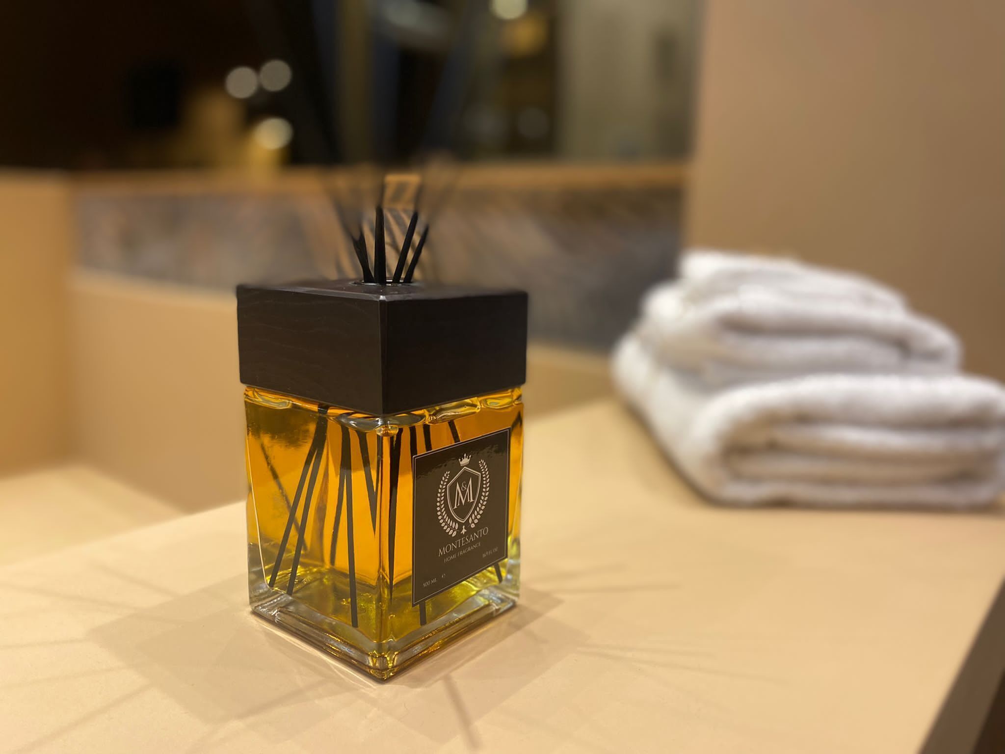 A square-shaped bottle of perfume with a black cap and a visible logo, placed on a table with a folded white towel in the background.