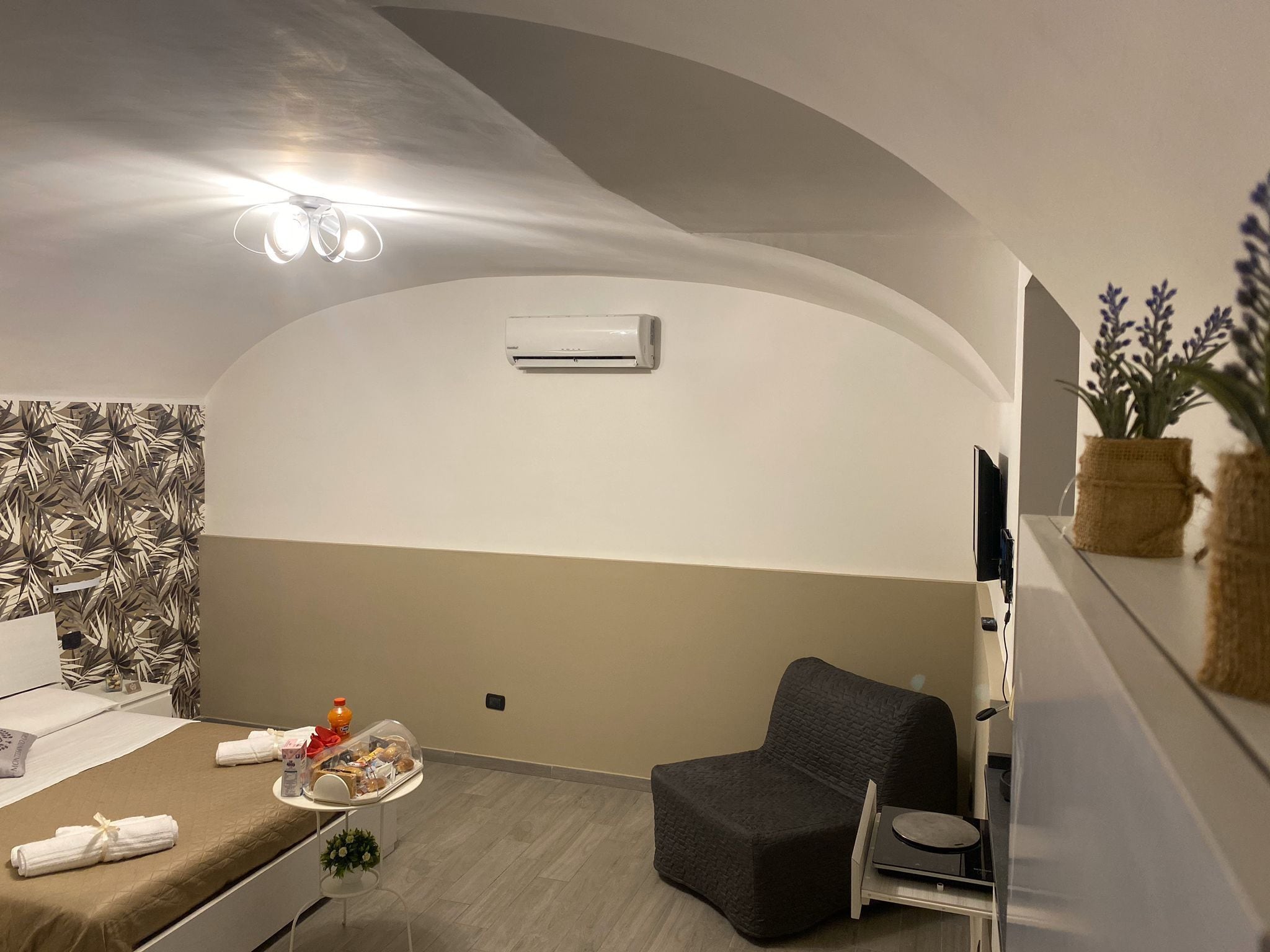 A hotel room with a bed, a small table with towels and snacks, a chair, a TV, and an air conditioning unit.