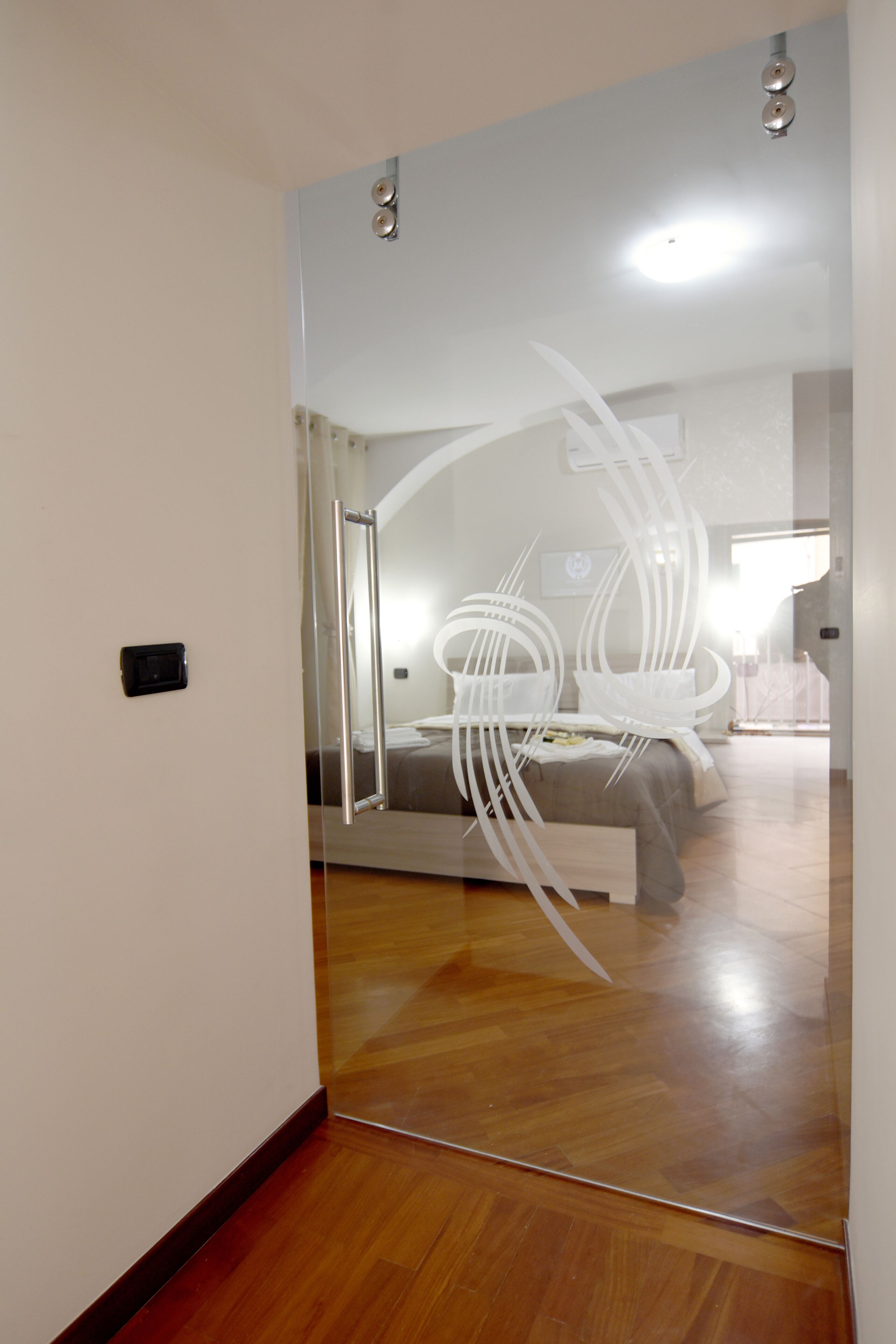 A modern hotel room with a wooden floor, a bed with a grey headboard, and a large mirror on the wall.