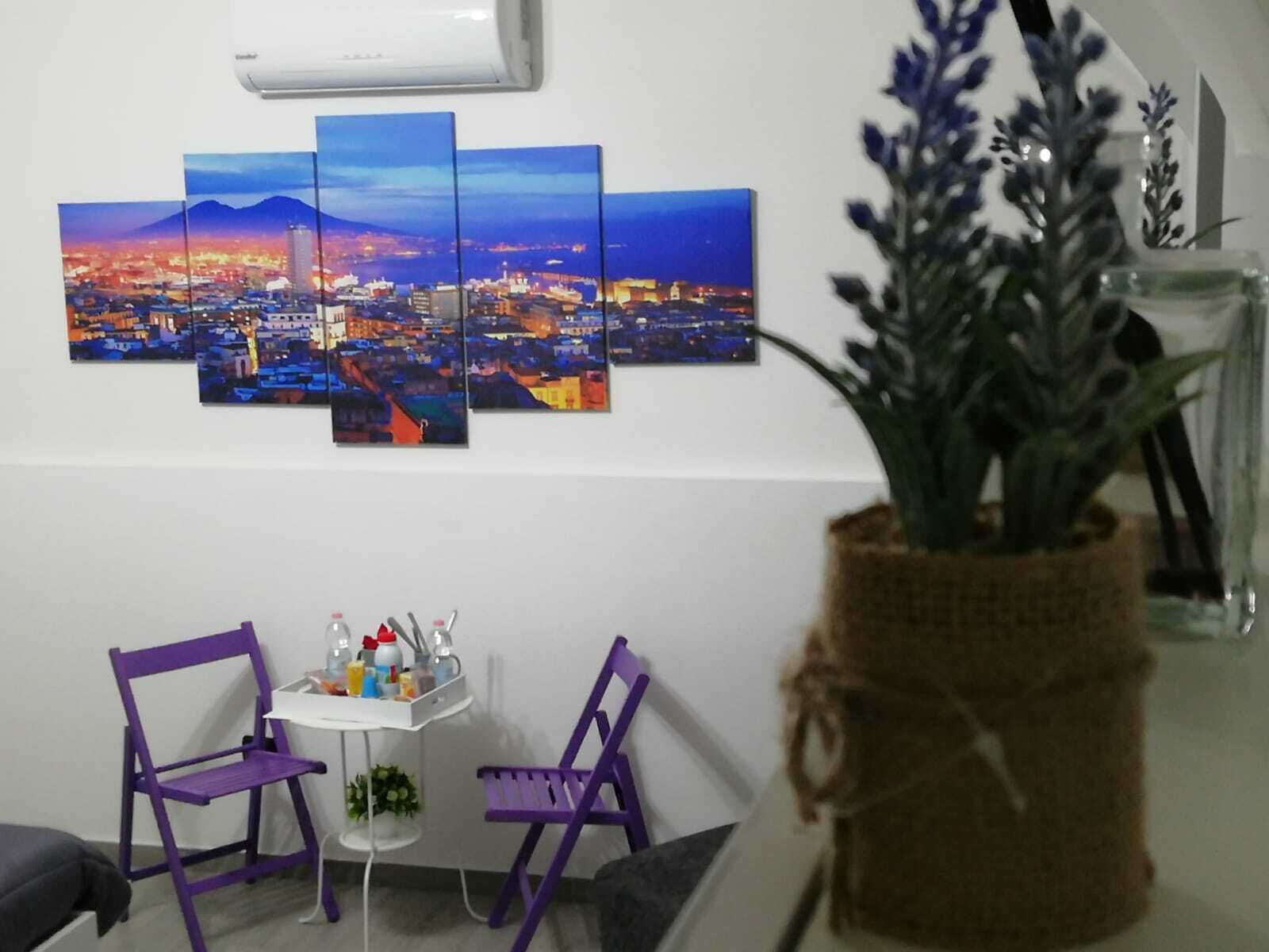 Ein Zimmer mit Stadtbildgemälde, zwei violette Stühle, einem kleinen Tisch mit Toilettenartikeln und einer Topfpflanze.