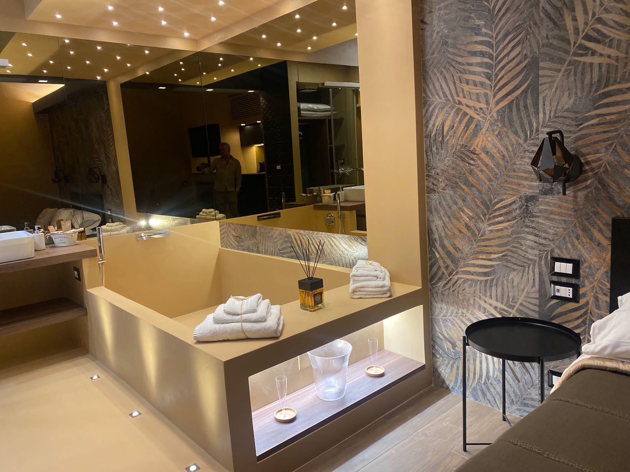 A modern bathroom with a large mirror, a bathtub, and a vanity area. There is a towel rack with folded towels and a small table with candles.