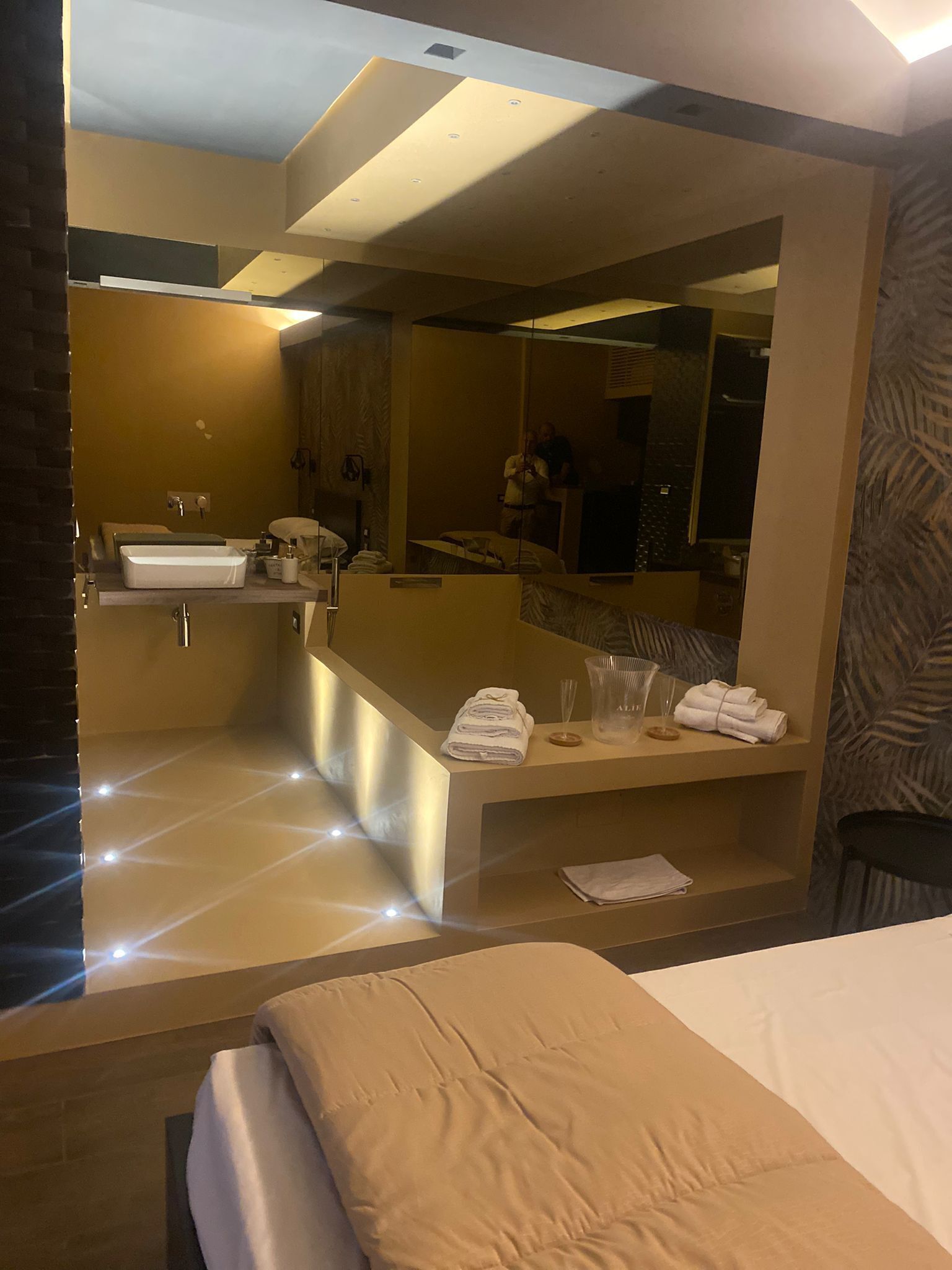 A modern hotel room with a large bed, a bathroom with a sink and mirror, and a small seating area with towels.