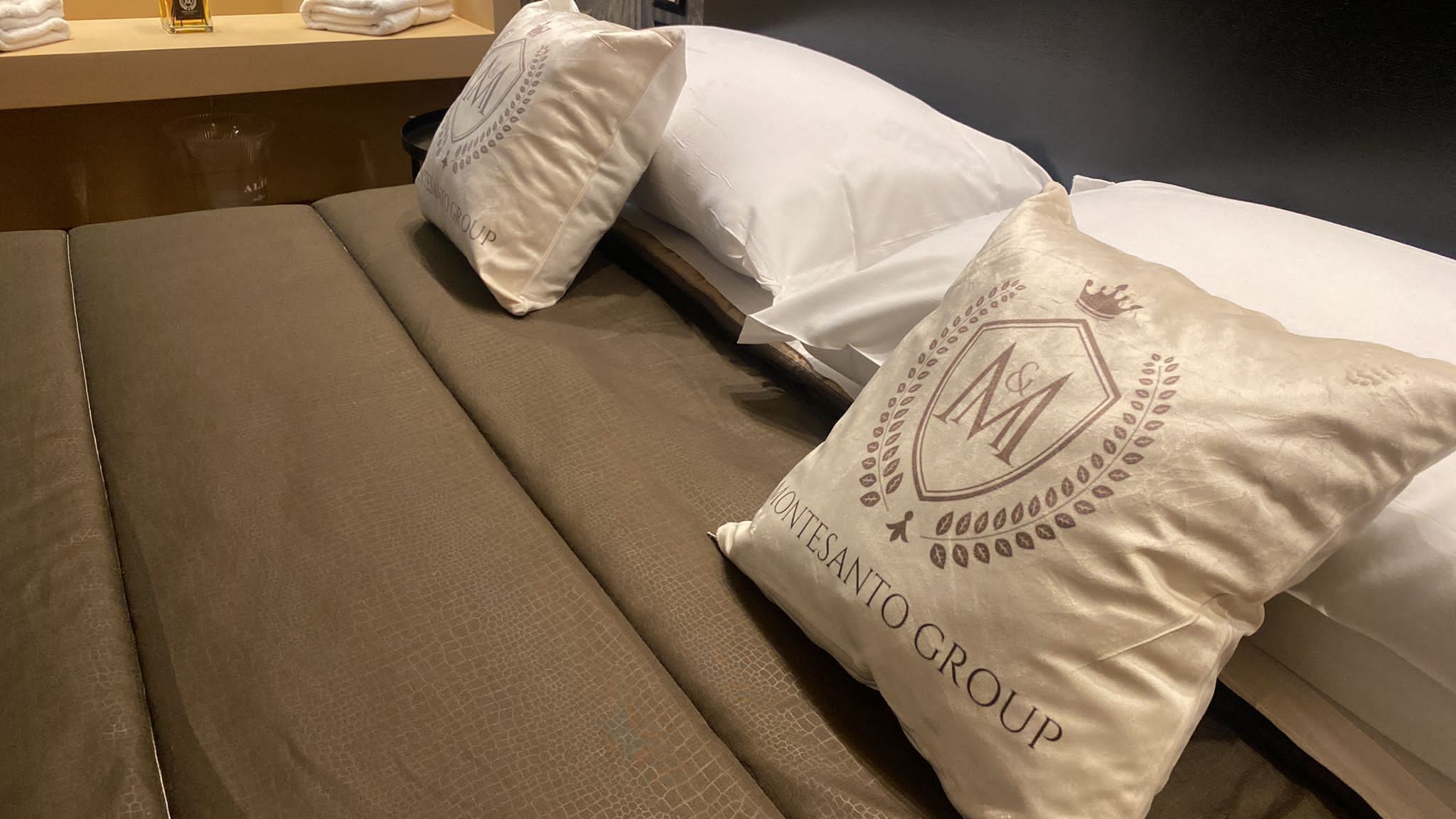 A hotel room with a bed featuring white linens and two decorative pillows with the logo "MONTESANTO GROUP".