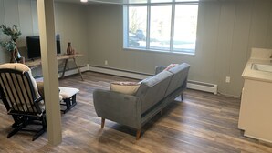A spacious living room with a grey sofa, a black chair, and a small table with decorative items. A large window provides natural light and a view of the outdoors. - Conneaut Creek Cottage (Conneaut)