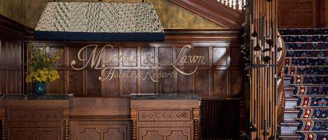 A grand lobby with a wooden reception desk, a large chandelier, and a staircase with ornate railings.