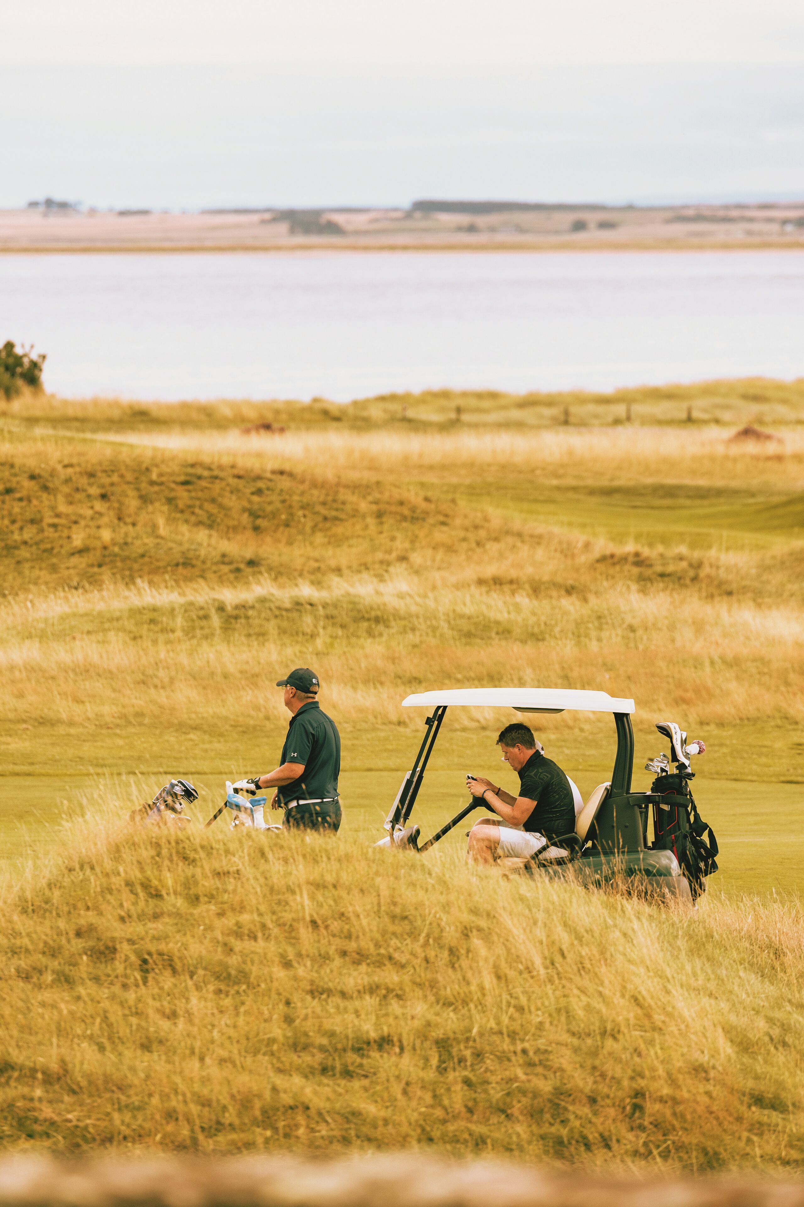 Campo de golf con un carrito y dos personas, una de pie y otra sentada, en un área verde con un cuerpo de agua al fondo.
