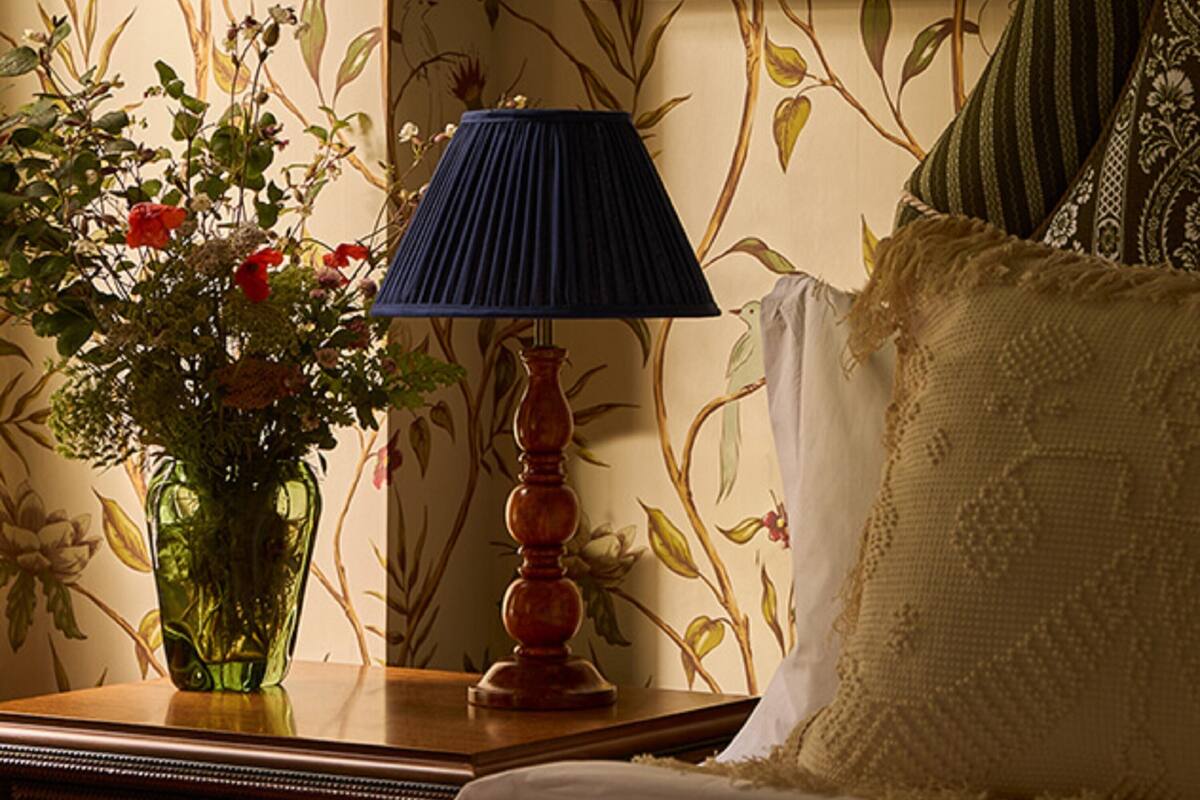 Une chambre à coucher avec un papier peint à motifs floraux, un tableau encadré au mur, une table de chevet avec une lampe et un lit recouvert d’une couverture bleue.