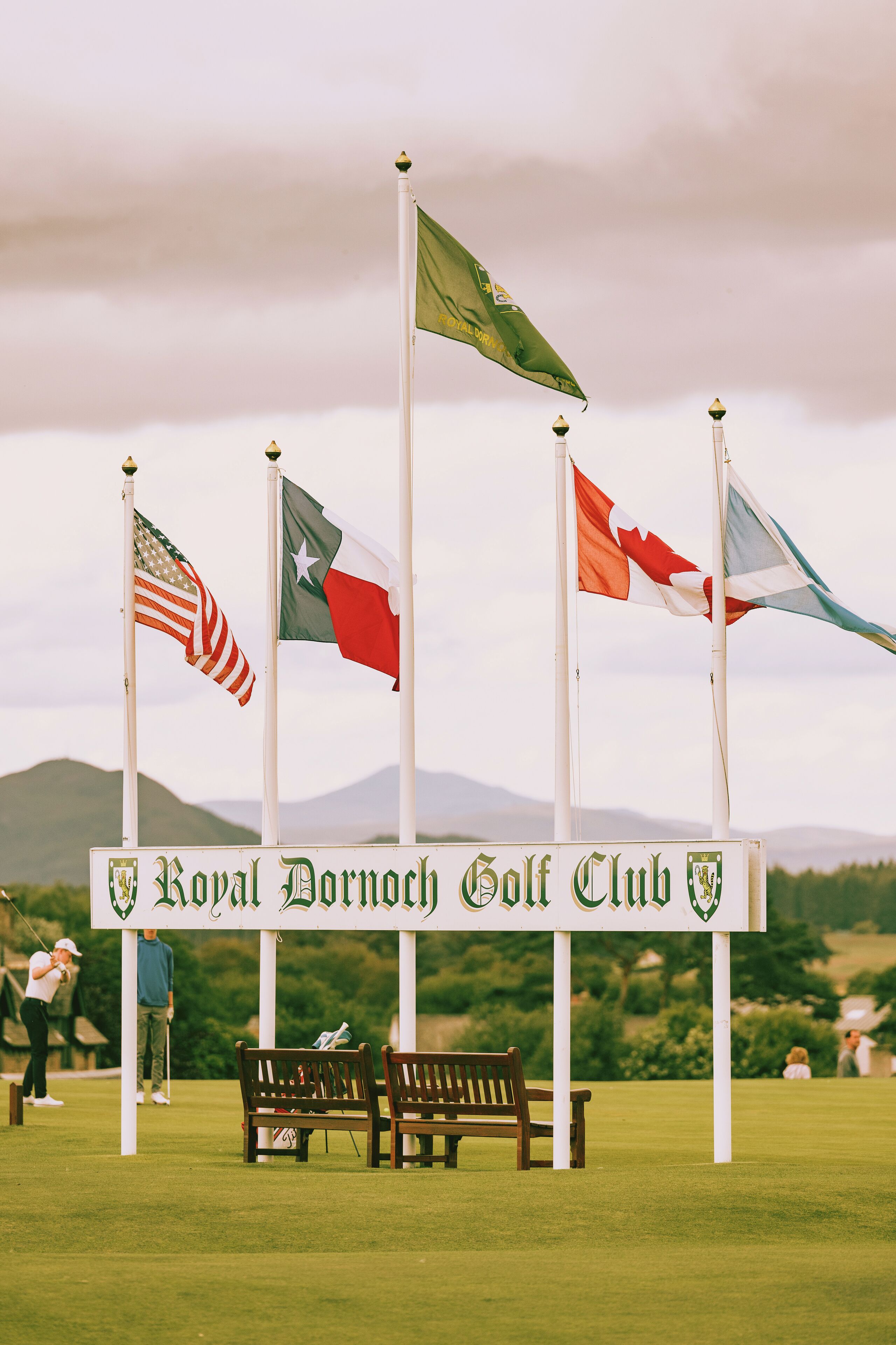 A golf club sign with multiple flags, a bench, and a mountainous background.