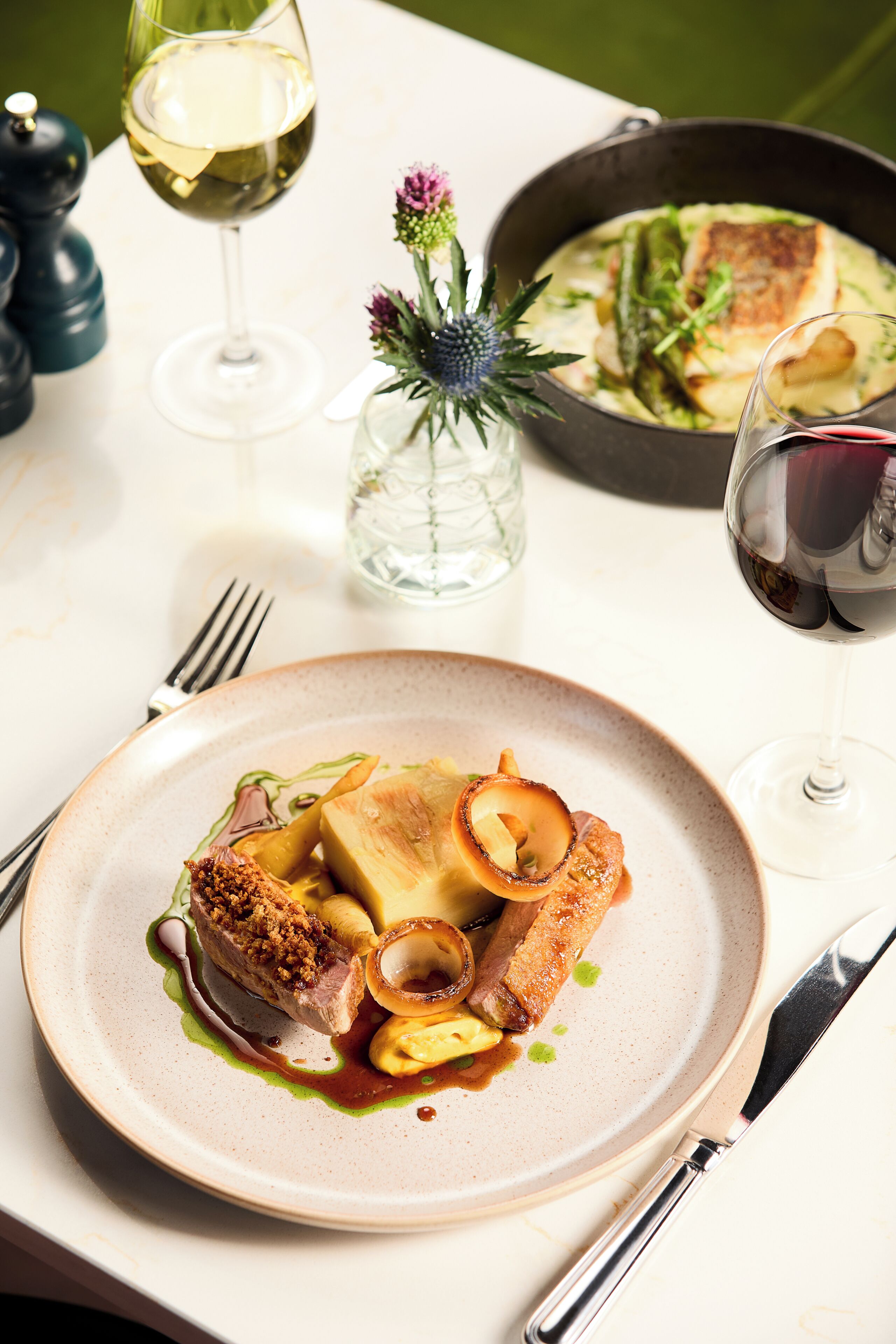 A dining table with a plate of food, a glass of white wine, and a pan with another dish.