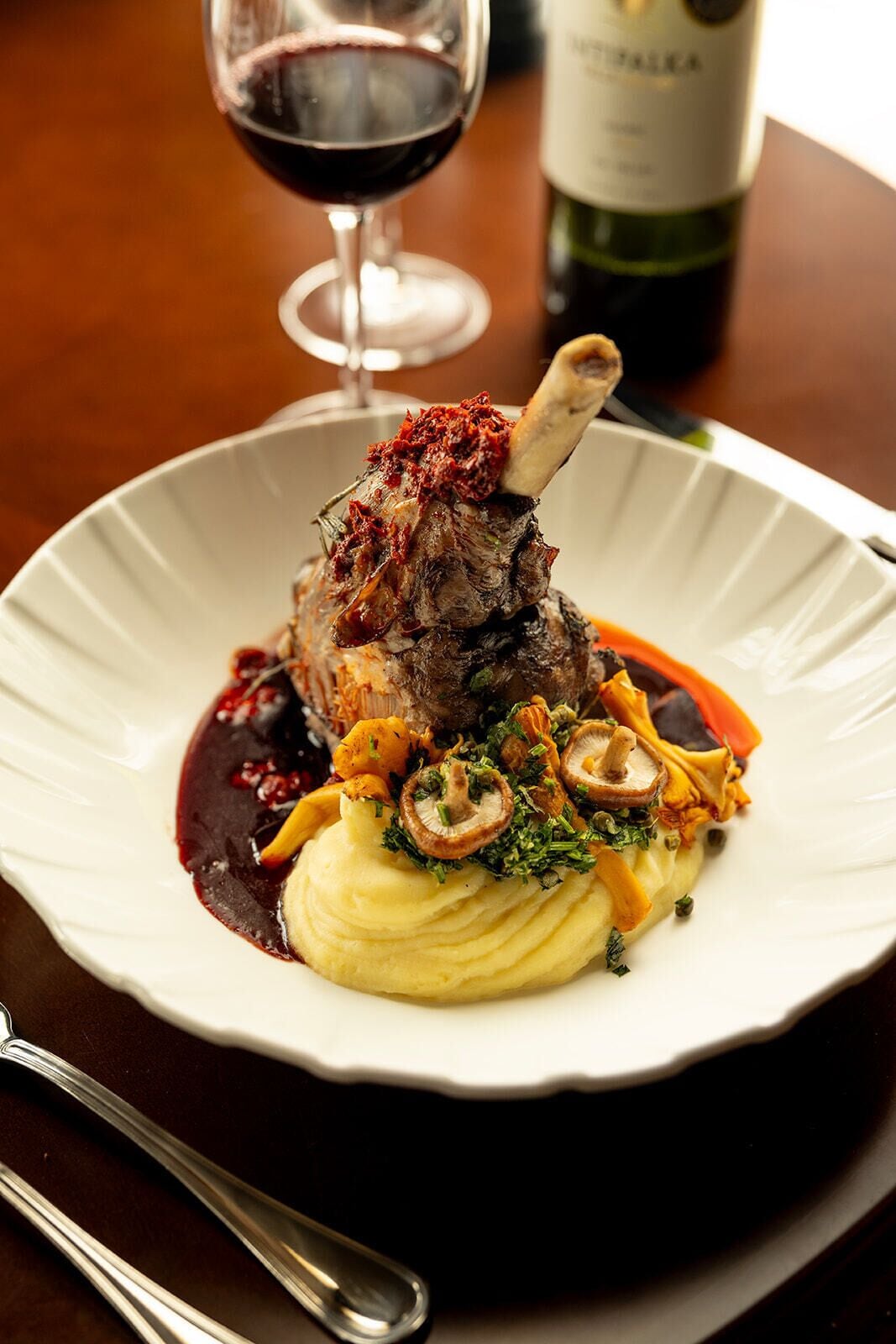 Une assiette contenant un plat de jarret d’agneau, de la purée de pommes de terre, des champignons et une sauce rouge.
