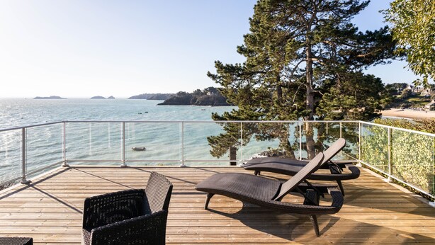 Terraza de madera con sillones de descanso y barandilla de vidrio con vista a una zona costera de aguas cristalinas y formas de tierra a lo lejos.