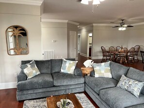 A living room with a grey sectional sofa, a wooden coffee table, and a dining area with wooden chairs and a table. - Waterfront Paradise (Bay St. Louis)
