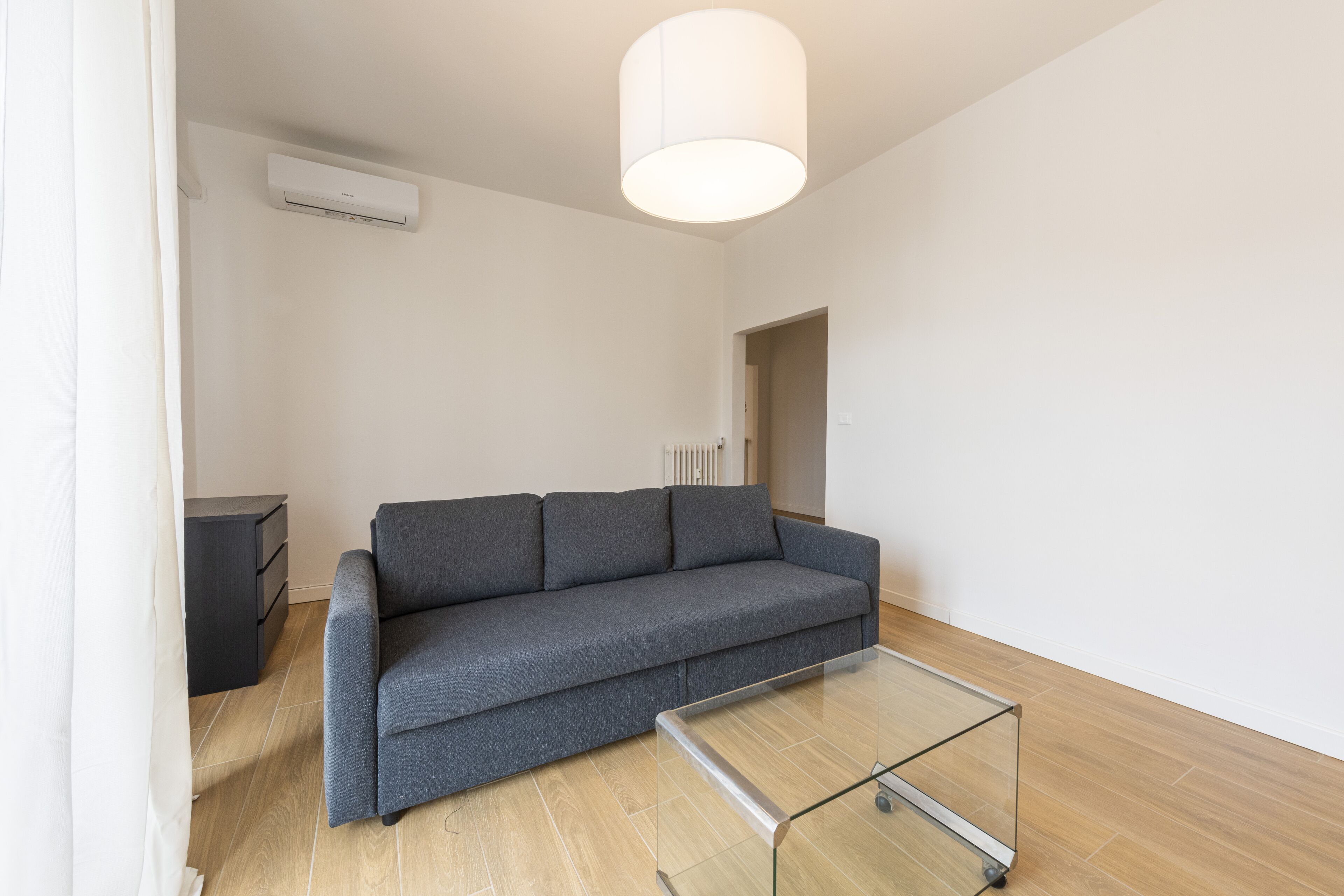 Una sala de estar moderna con un sofá gris, una mesa de centro de vidrio, una lámpara de techo blanca y un radiador.