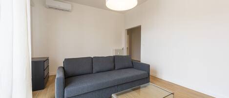 Una sala de estar moderna con un sofá gris, una mesa de centro de vidrio, una lámpara de techo blanca y un radiador.