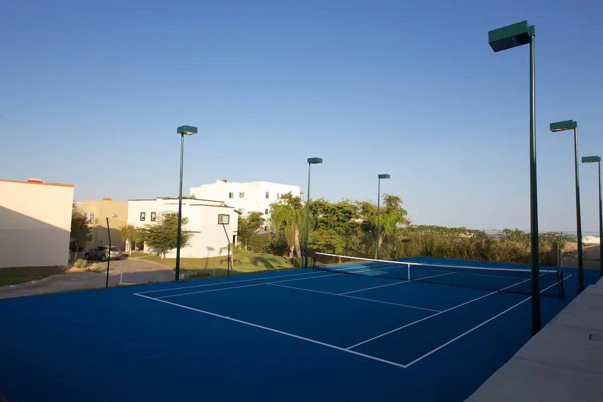 An outdoor tennis court with a blue surface, surrounded by buildings and trees.