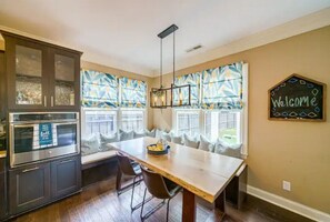 A kitchen with a built-in oven, a dining table with chairs, and large windows with patterned curtains. - South End Home: Pool + Spa+ Firepit+ Arcade (Charlotte)