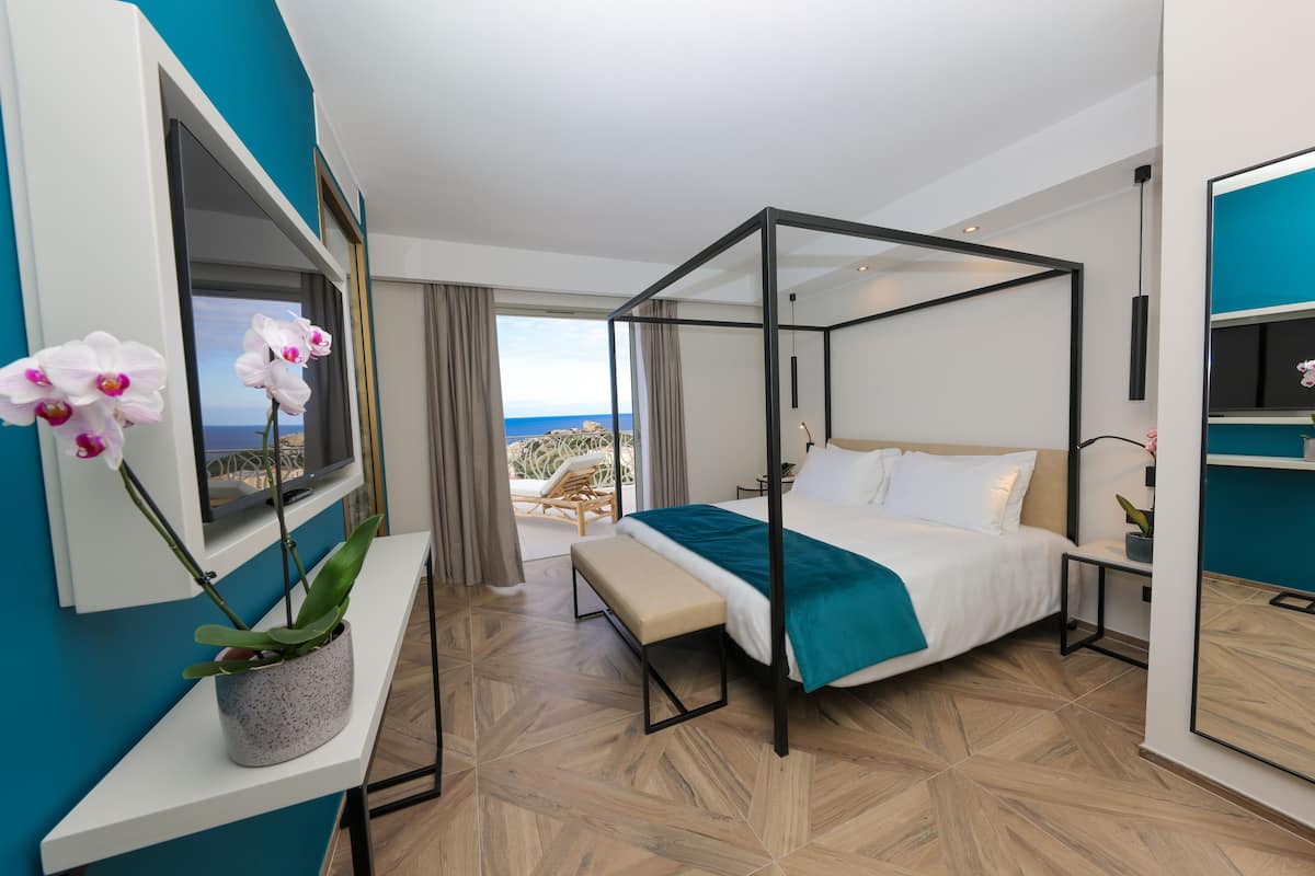 Quarto de hotel moderno com cama de dossel, um banco, uma orquídea em vaso e vista para o oceano.