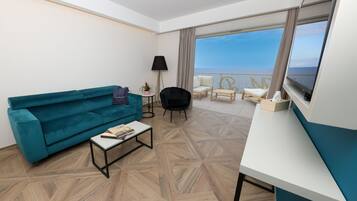Habitación de hotel moderna con sofá color turquesa, una pequeña mesa de centro, televisor y balcón con vista al mar.