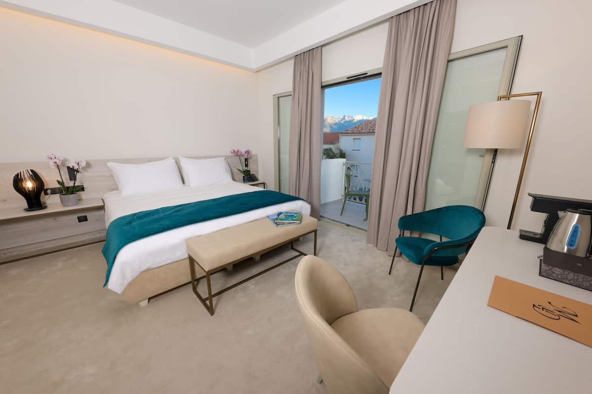 Quarto de hotel com cama, escrivaninha, cadeira, luminária e vista para montanhas pela janela.