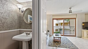 A bathroom with a sink, mirror, and wall sconces. A sliding glass door leads to a balcony with a TV and a small table. - Well-appointed condo on uncrowded Ormond Beach (Ormond Beach)
