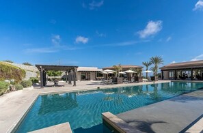 A swimming pool with lounge chairs and umbrellas, surrounded by buildings and palm trees. - Departamentos TT2 (Cabo San Lucas)