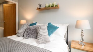 A neatly made bed with a grey blanket and multiple pillows, a bedside table with a lamp, and a wooden shelf with candles and greenery. - Ferry View (Poulton-le-Fylde)