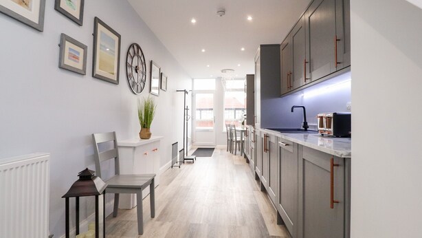A modern kitchen with grey cabinets, a white countertop, and a dining area with a chair and a small table. - Ferry View (Poulton-le-Fylde)