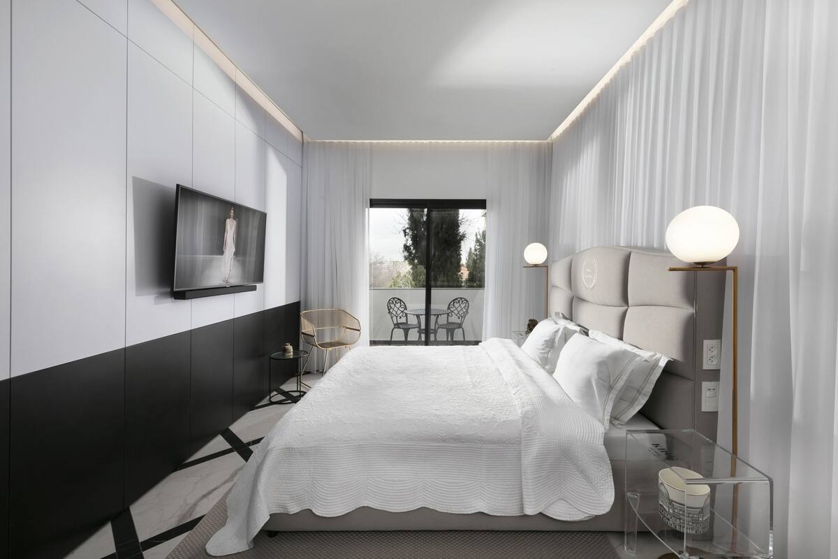 Une chambre moderne avec un grand lit, un téléviseur fixé au mur, un balcon avec des chaises et une table de chevet avec une lampe.