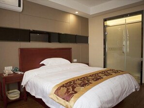 Quarto de hotel com uma cama grande, uma mesa de cabeceira de madeira com telefone, chuveiro com divisória de vidro e uma televisão na parede.